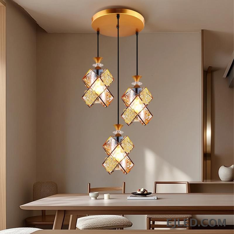 This three-light pendant fixture features gold-finished frames and clear glass panels forming distinct, double-box shades. Each shade encloses a single lamp, providing warm illumination from a round gold ceiling plate. - view 3