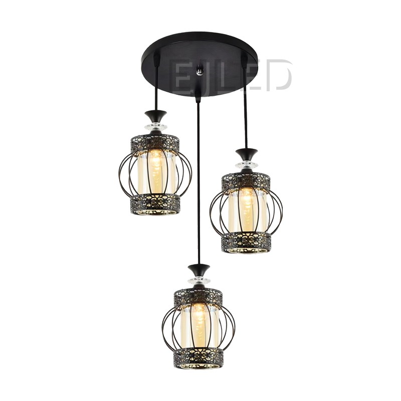 Esta lámpara colgante de hierro negro con tres luces presenta jaulas cilíndricas abiertas con detalladas bandas metálicas y pantallas interiores de cristal ámbar, suspendidas de un dosel redondo de techo.