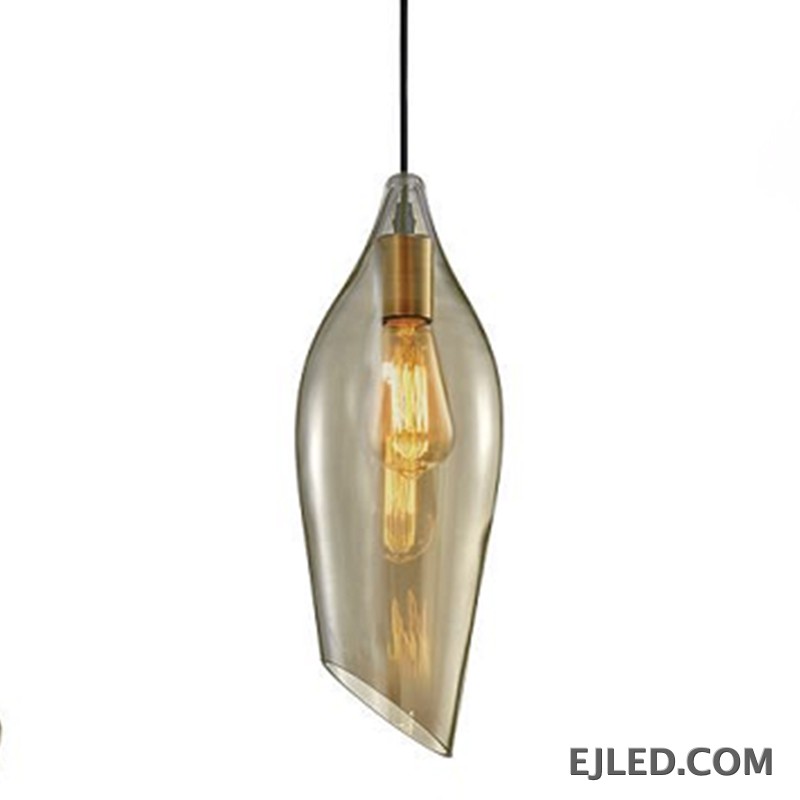 This glass pendant light features a unique, asymmetrical leaf-like shade in a warm amber tint, housing a single visible lamp with a brass-toned socket. It hangs from a simple black cord, offering an open bottom for light dispersal.