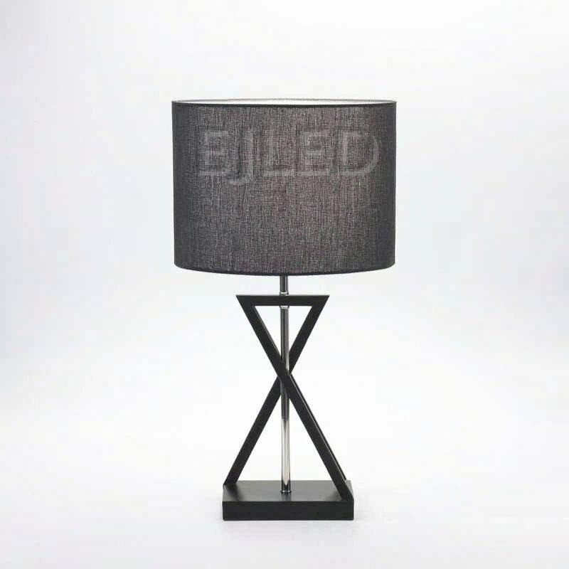This black fabric shade table lamp features an hourglass geometric metal base in a dark finish and a central chrome support rod, blending modern lines with soft illumination for indoor spaces.