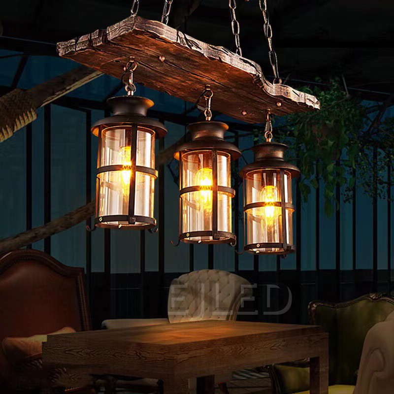 This wooden linear pendant lighting features three cylindrical lantern-style lamps suspended from a distressed wood beam. It hangs via chains from a dark metal ceiling plate, offering a rustic aesthetic. - view 3