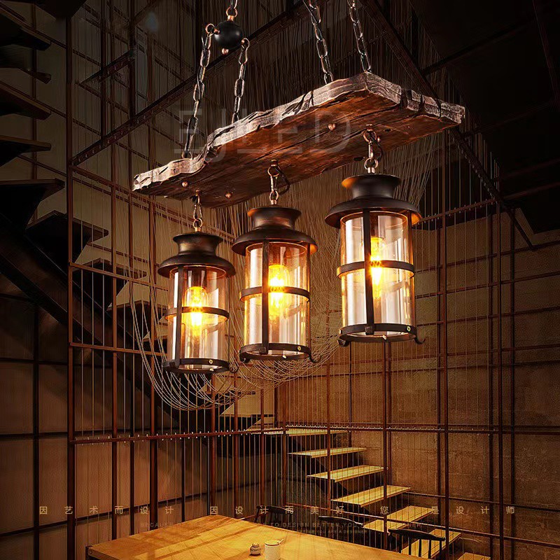 This wooden linear pendant lighting features three cylindrical lantern-style lamps suspended from a distressed wood beam. It hangs via chains from a dark metal ceiling plate, offering a rustic aesthetic. - view 5