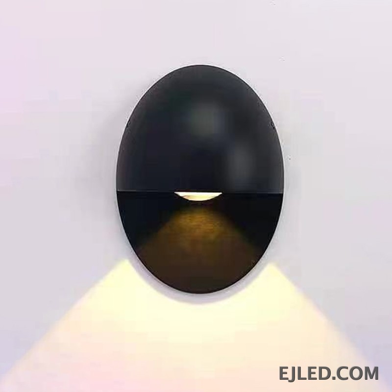 This black oval aluminum wall sconce projects a soft, downward light. Its distinctive design features a concealed light source within the lower half, providing indirect illumination. - view 2