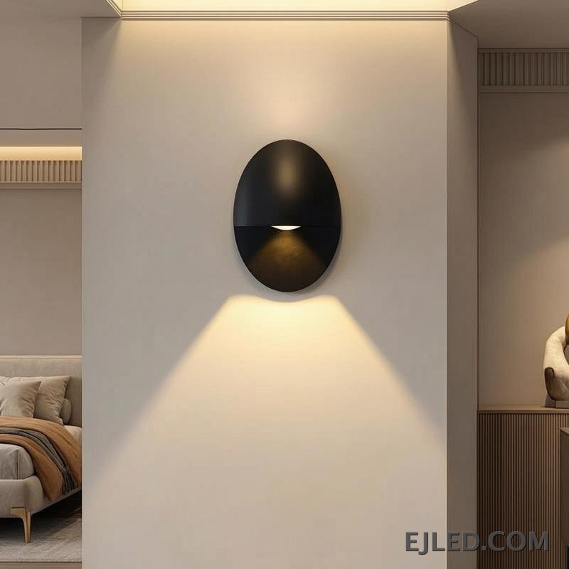 This black oval aluminum wall sconce projects a soft, downward light. Its distinctive design features a concealed light source within the lower half, providing indirect illumination. - view 4