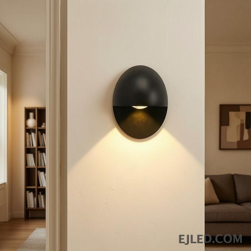 This black oval aluminum wall sconce projects a soft, downward light. Its distinctive design features a concealed light source within the lower half, providing indirect illumination. - view 5