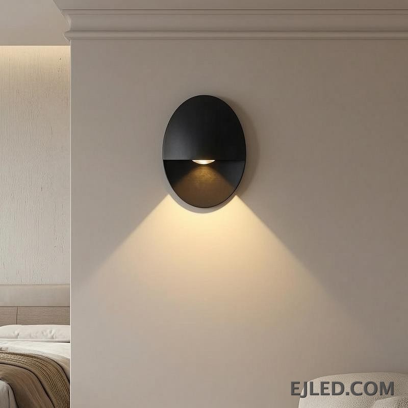 This black oval aluminum wall sconce projects a soft, downward light. Its distinctive design features a concealed light source within the lower half, providing indirect illumination. - view 6